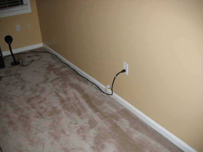 Corner of a room with tan walls, carpet, and a black power cord plugged into a wall outlet