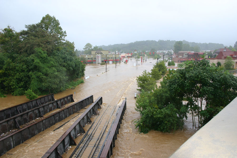 Flooded street with buildings and trees, railway bridge in foreground
