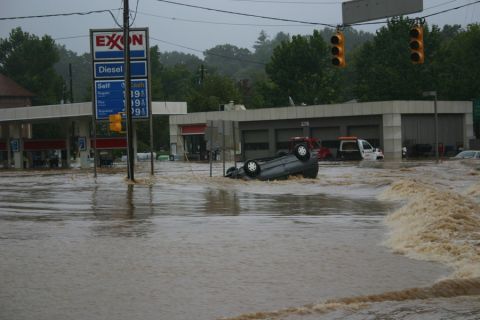 1985 Ford LTD Crown Victoria - A car is overturned in floodwaters near an Exxon gas station, with other vehicles visible in the background.
