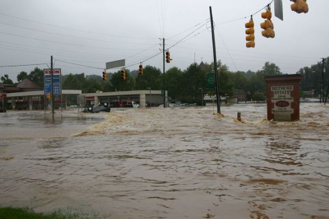 1985 Ford LTD Crown Victoria - A car is overturned in a flooded street, with a gas station and a brick sign visible.