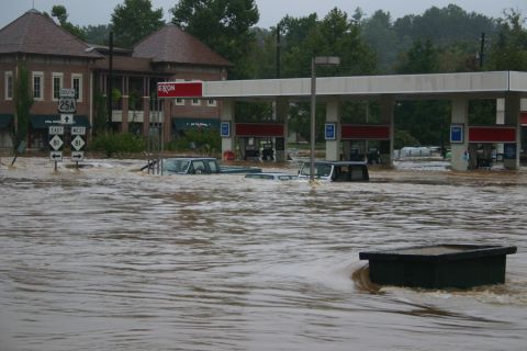 1985 Ford LTD Crown Victoria - Several vehicles are submerged in floodwaters near a gas station, with buildings and trees visible in the background.