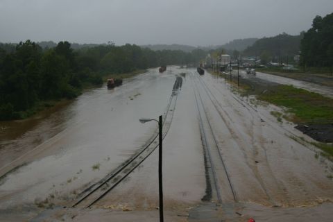 1985 Ford LTD Crown Victoria - Train tracks and a road are submerged in floodwaters, with trucks navigating the high water.
