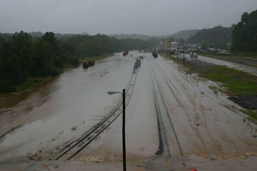 Flooded train tracks and a road with several trucks visible during a heavy rain event