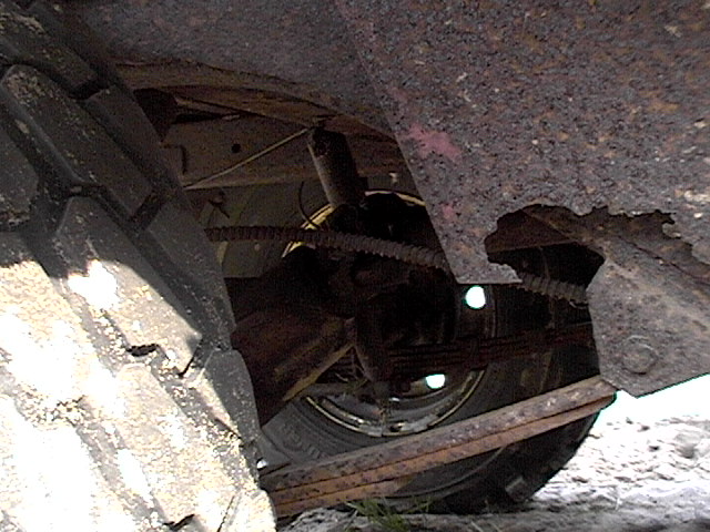 Underside of a vehicle showing rusty leaf springs, shock, drum brake, and a knobby tire, with snow on the ground