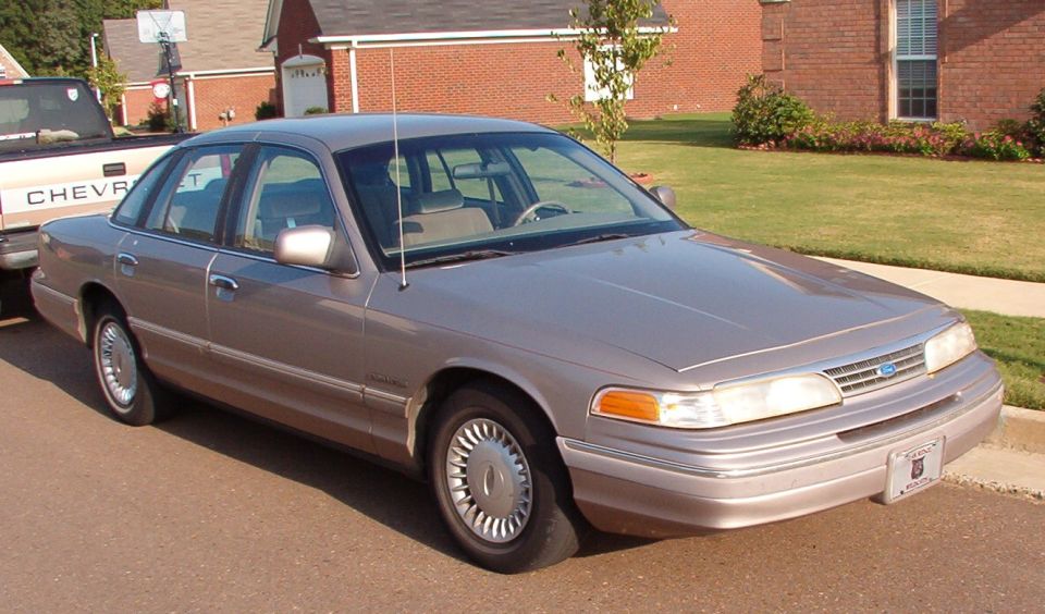 Silver Ford Crown Victoria sedan, front three-quarter view, parked by a brick house.