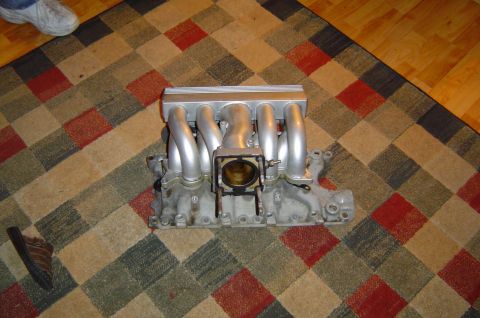 1993 Ford Bronco - An intake manifold is shown on a patterned rug, with a shoe and a person's leg visible in the background.
