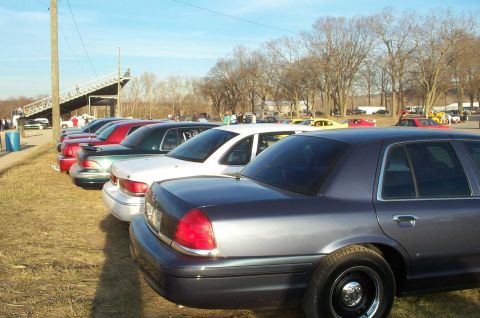1998 Ford Crown Victoria - Several sedans are parked in a lot near a grandstand, with trees in the background.
