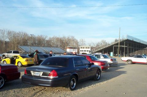 1998 Ford Crown Victoria - Several cars are parked at a raceway, with grandstands visible in the background under a clear sky.
