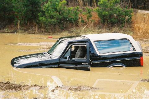 1995 Ford Bronco - A black and white Ford Bronco is submerged in muddy water with its driver's door ajar.