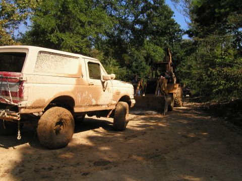 1989 Ford Bronco - A muddy 1989 Ford Bronco is parked on a dirt path, with a backhoe visible behind it in a wooded area.