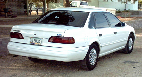 White Ford Taurus sedan rear three-quarter view with U-SUCK license plate, parked on gravel