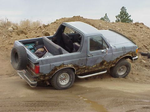 1989 Ford Bronco - Grey-blue full-size Ford Bronco with the rear top removed, flexing its suspension over a dirt mound off-road, seen from the rear three-quar…
