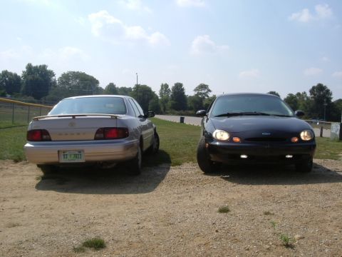 1996 Ford Taurus - Two sedans, a black Ford and a tan Toyota, are parked on a dirt lot with a baseball field in the background.