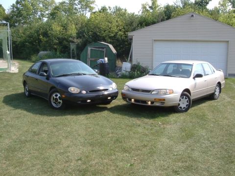 1996 Ford Taurus - Two sedans are parked on a grassy area in front of a beige garage, with trees and a small shed in the background.