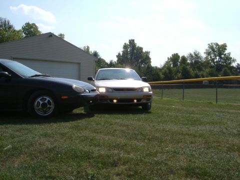 1996 Ford Taurus - Two sedans, one a 1996 Ford Taurus, are parked on the grass near a garage and a baseball field.