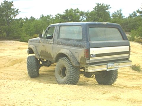 1995 Ford Bronco - Rear three-quarter view of a lifted two-tone blue-over-silver 1990s Ford Bronco parked on packed sand, showing the hardtop, large rear glas…