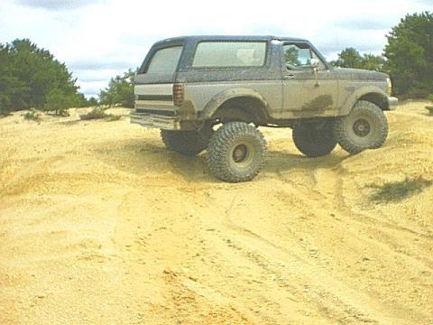 1995 Ford Bronco - Side view of a lifted two-tone 1990s Ford Bronco with a removable hardtop, straddling a sand berm in an open sand pit with the rear tire li…