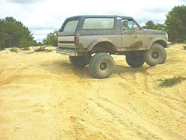 Lifted two-tone 90s Ford Bronco climbing a sand berm on mud tires, side view in a sand pit