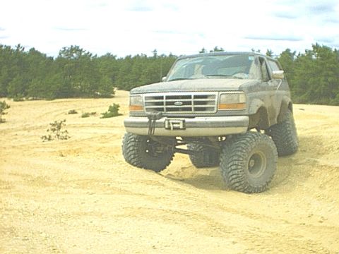 1995 Ford Bronco - A lifted 1990s Ford Bronco sits in an open sand pit under a bright hazy sky, front three-quarter view showing tall mud-terrain tires on ste…