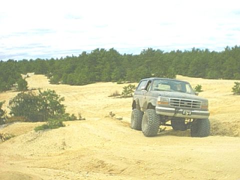 1995 Ford Bronco - A lifted 1990s Ford Bronco on tall mud-terrain tires sits atop a sand ridge in a wide open sand pit ringed by pine forest, seen from a low…