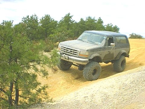 1995 Ford Bronco - A lifted 1990s Ford Bronco with a hardtop descends a sandy bank in a pine-lined sand pit, front three-quarter view on tall mud-terrain tire…