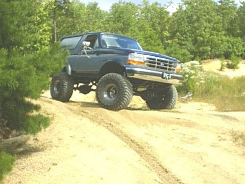 1995 Ford Bronco - A lifted blue 1990s Ford Bronco with a hardtop crests a sandy trail berm, front wheels flexed on tall mud-terrain tires and steel wheels. T…