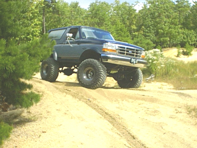 Lifted blue 90s Ford Bronco on tall mud tires climbing a sandy trail berm in a pine sand pit
