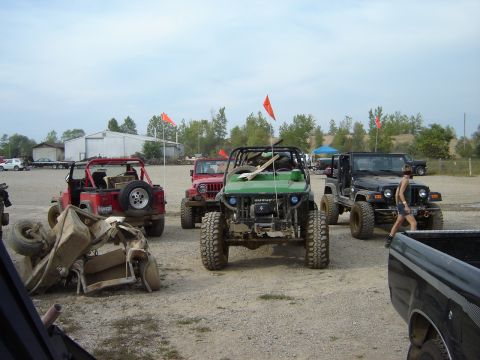 1992 Ford Bronco - A group of modified off-road vehicles are gathered in an outdoor lot, with one vehicle flipped on its side.