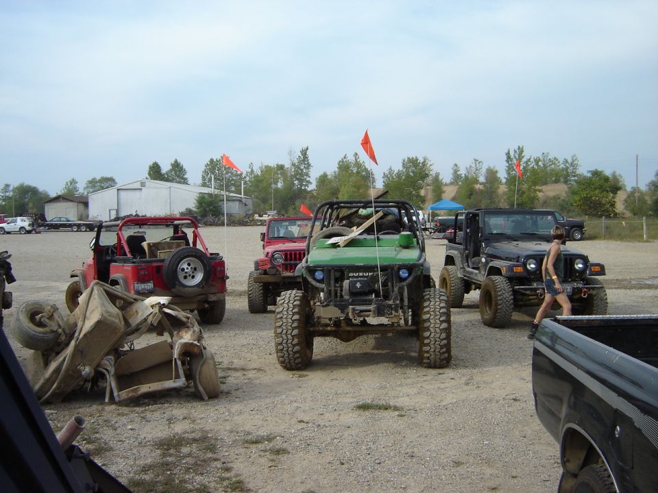 Several off-road vehicles, including a green Suzuki Samurai and red Jeeps, parked in a dirt lot. One Jeep is overturned