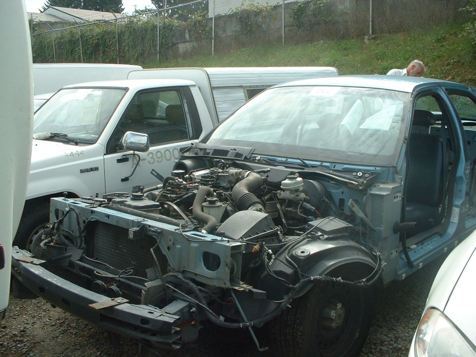 Light blue sedan with front end removed, engine exposed. A white pickup truck is to the left