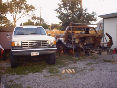 1988 Ford Bronco - A white 1987 Ford F-Series truck is parked next to a severely damaged vehicle frame in a yard.