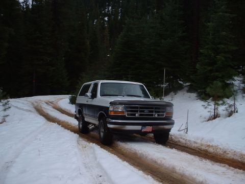 1993 Ford Bronco - A two-tone 1993 Ford Bronco navigates a snowy, rutted track through a forest.