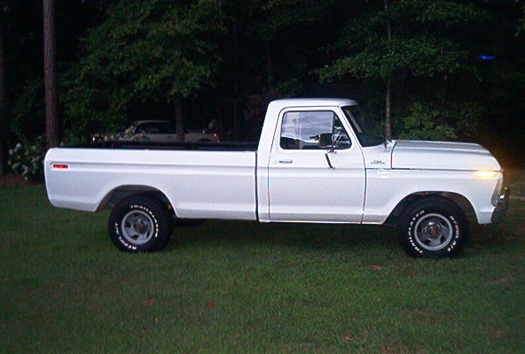 White vintage Ford pickup truck, side view, parked on grass in front of trees at dusk