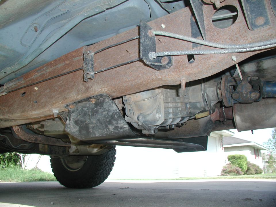 Underside view of vehicle frame, transmission, driveshaft, and exhaust system. Tire visible