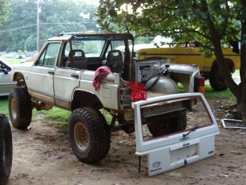 1985 Ford Bronco - A white Jeep Comanche pickup truck sits disassembled in an outdoor setting, with a yellow truck visible in the background.
