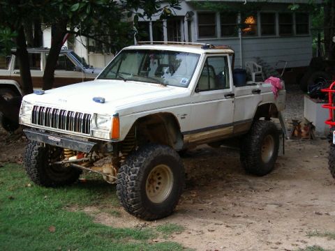 1985 Ford Bronco - A lifted white Jeep Comanche pickup truck is shown with large off-road tires and modifications, parked outdoors.