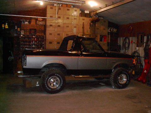1989 Ford Bronco - Side view of a two-tone black and silver Ford Bronco with aftermarket wheels and tires, parked inside a garage.