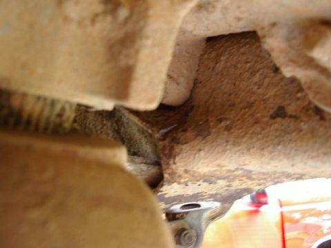 1985 Ford Bronco - Close-up underbody shot showing heavily rusted and mud-caked frame rail and sheet metal, with a corroded threaded rod or coil spring visibl…