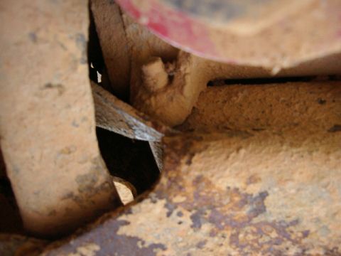 1985 Ford Bronco - Tight underbody close-up of a dirt-caked, surface-rusted frame rail and bracket with a torn or cut sheet-metal edge and a bolt stub protrud…