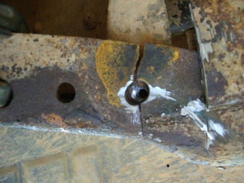1985 Ford Bronco - Close-up of a heavily rusted boxed steel frame rail showing a crack propagating downward from a bolt hole, with white chalk or paint marks…