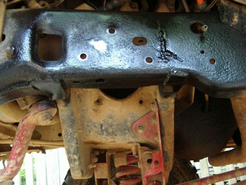 1985 Ford Bronco - Underside view of a freshly painted glossy black frame crossmember with fresh weld beads and drilled mounting holes, spanning above a dirt-…