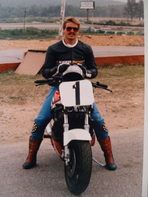 1985 Ford LTD Crown Victoria - A motorcycle racer sits on a bike with a number 1 plate, dressed in a racing suit and helmet.