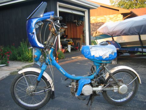 1985 Ford LTD Crown Victoria - A blue Yamaha moped is parked in front of a garage with a boat on a trailer behind it.