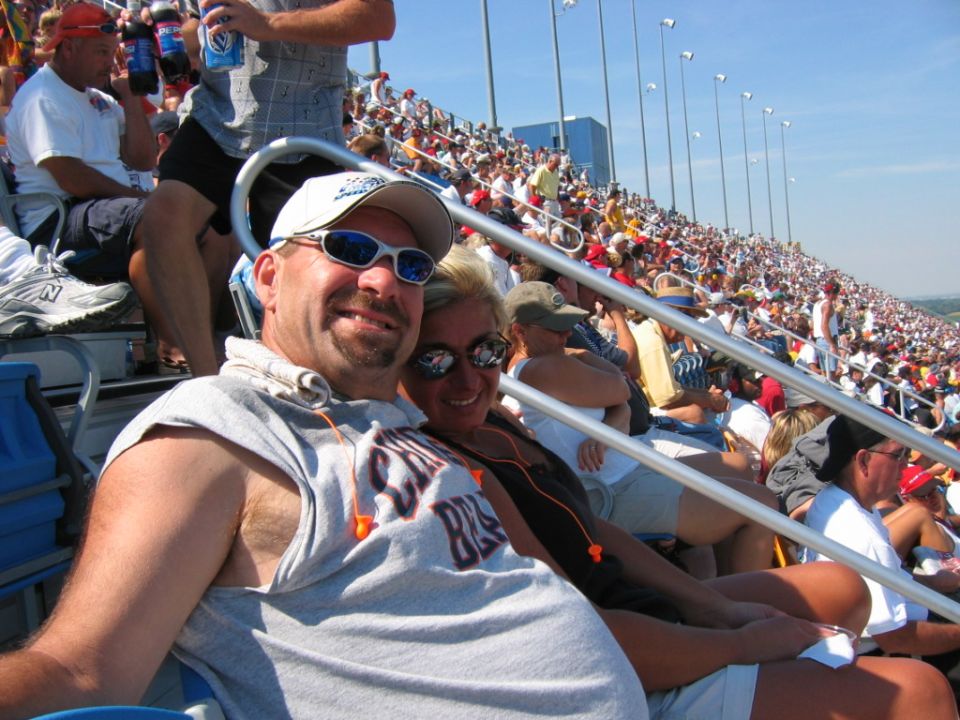 Two people smile for a selfie in stadium seats with a large crowd in the background