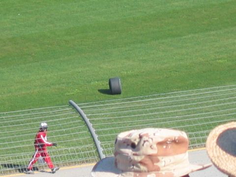1985 Ford LTD Crown Victoria - A lone racing tire rests on the grass beside a fence, with a person in a red racing suit walking nearby.