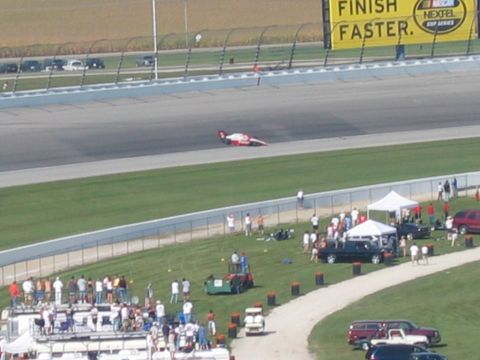 1985 Ford LTD Crown Victoria - A race car speeds around the track, with a crowd gathered in the infield and tents set up nearby.