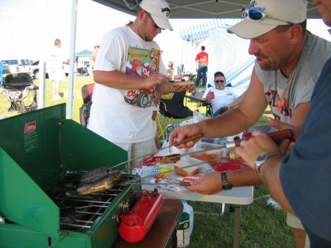 1985 Ford LTD Crown Victoria - Outdoor cooking scene with multiple people preparing food on a portable grill.