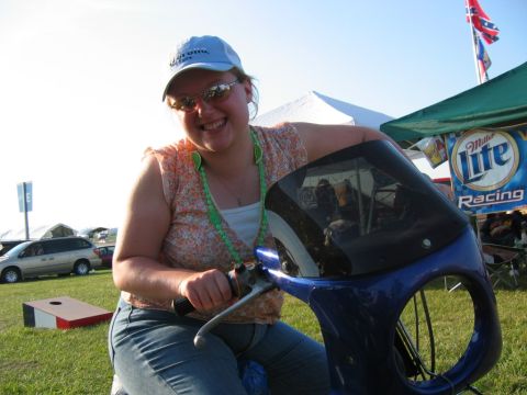 1985 Ford LTD Crown Victoria - A woman sits on a blue scooter with a black windshield, with tents and a van in the background.