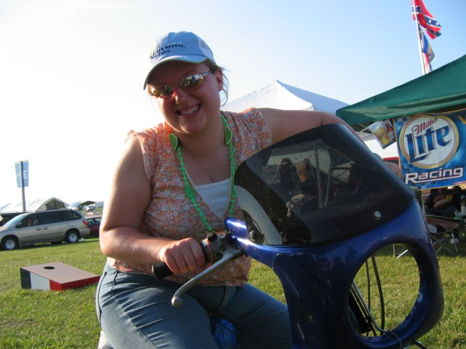 Woman on a blue scooter with a black windshield, wearing a Corona hat and sunglasses