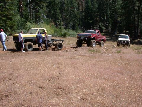 1996 Ford Bronco - A yellow and grey pickup truck chassis sits in a field, with two other trucks visible in the background.
